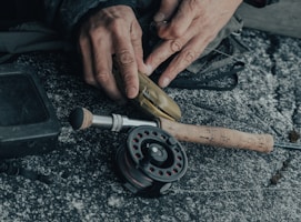 A fisherman's hands tying a hook with fishing line near a calm lake at dawn.