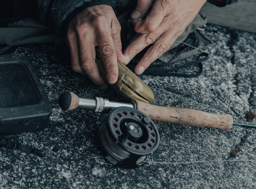 Hands are seen threading fishing line onto a fishing reel. The reel is attached to a rod with a wooden handle, placed on a textured surface. A square black box is visible beside the rod.