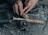 Hands are seen threading fishing line onto a fishing reel. The reel is attached to a rod with a wooden handle, placed on a textured surface. A square black box is visible beside the rod.