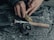 Hands are seen threading fishing line onto a fishing reel. The reel is attached to a rod with a wooden handle, placed on a textured surface. A square black box is visible beside the rod.