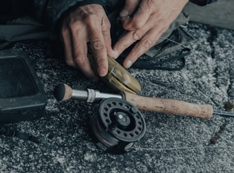 Hands are seen threading fishing line onto a fishing reel. The reel is attached to a rod with a wooden handle, placed on a textured surface. A square black box is visible beside the rod.