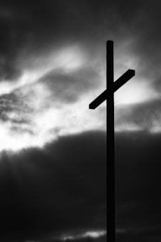 A silhouette of a cross stands against a backdrop of dark, dramatic clouds with light breaking through, creating a stark and powerful visual contrast.