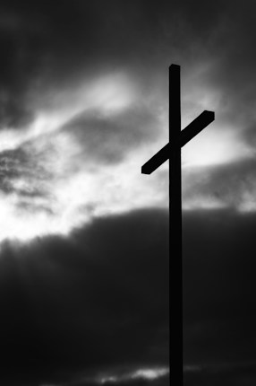 A silhouette of a cross stands against a backdrop of dark, dramatic clouds with light breaking through, creating a stark and powerful visual contrast.
