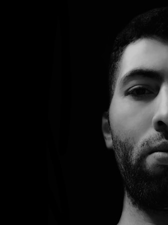 A close-up, black and white photograph of a man with facial hair, featuring just the right half of his face against a completely black background. The image highlights the details and contours of the forehead, eye, and part of the mouth, creating a dramatic contrast.
