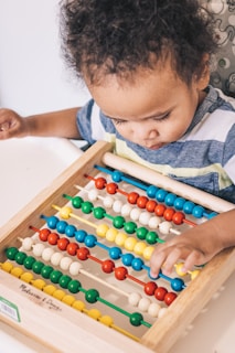 A young child is engaged with a colorful wooden abacus, focusing intently on the counting beads. The abacus features rows of alternating colors with beads in red, blue, green, yellow, and white. The child appears curious and concentrated as they manipulate the beads.