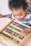 A young child is engaged with a colorful wooden abacus, focusing intently on the counting beads. The abacus features rows of alternating colors with beads in red, blue, green, yellow, and white. The child appears curious and concentrated as they manipulate the beads.