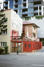 A small rustic building with a red exterior and wooden doors appears to be a café or bar titled 'Paladar Fumior Salon.' It features signs advertising espresso and Cuban cigars. The structure is situated on a city street corner with a modern multi-story building in the background. A 'No Entry' sign is visible next to the bar, and some greenery is present around the area.