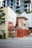 A small rustic building with a red exterior and wooden doors appears to be a café or bar titled 'Paladar Fumior Salon.' It features signs advertising espresso and Cuban cigars. The structure is situated on a city street corner with a modern multi-story building in the background. A 'No Entry' sign is visible next to the bar, and some greenery is present around the area.