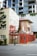 A small rustic building with a red exterior and wooden doors appears to be a café or bar titled 'Paladar Fumior Salon.' It features signs advertising espresso and Cuban cigars. The structure is situated on a city street corner with a modern multi-story building in the background. A 'No Entry' sign is visible next to the bar, and some greenery is present around the area.