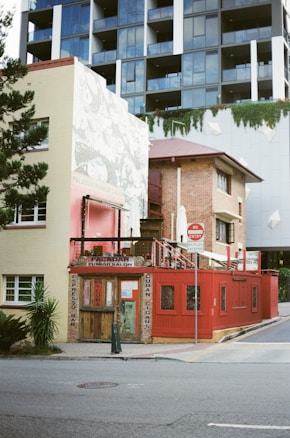 A small rustic building with a red exterior and wooden doors appears to be a café or bar titled 'Paladar Fumior Salon.' It features signs advertising espresso and Cuban cigars. The structure is situated on a city street corner with a modern multi-story building in the background. A 'No Entry' sign is visible next to the bar, and some greenery is present around the area.