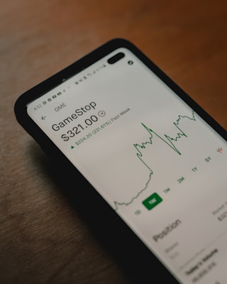 A smartphone screen displays stock information for GameStop, showing a price of $321.00 with a notable increase in value over the past week. The screen includes a graph depicting the stock's recent performance.