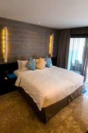 A neatly made bed with a white duvet and multiple decorative pillows is placed against a dark wood accent wall. The pillows include two with blue and white patterns and a central beige pillow. Elegant wall sconces with soft lighting are mounted on either side, adding warmth. Heavy curtains partially cover a large window, allowing some natural light to enter the room.