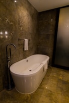A modern bathroom featuring a sleek, freestanding bathtub with a curved design. The walls and floor have a marble finish in various shades of brown, enhancing the luxurious feel. A white towel is draped over the side of the tub, and there is a polished silver faucet nearby. The ambient lighting casts soft reflections on the marble surfaces.