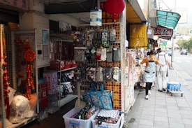 A busy street market with various items displayed for sale, including gloves, socks, decorative ornaments, and bags. A woman wearing an apron stands beside the shop entrance while another person walks away pushing a cart.
