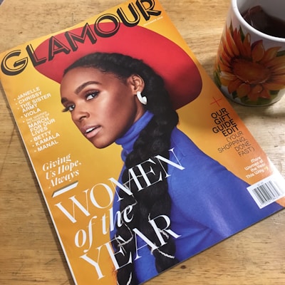 A magazine titled 'Glamour' featuring a stylish woman with a braided hairstyle, wearing a red hat and a blue outfit. It highlights the 'Women of the Year' issue and includes other notable names. A cup of tea with a sunflower design is placed beside the magazine on a wooden surface.
