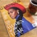 A magazine titled 'Glamour' featuring a stylish woman with a braided hairstyle, wearing a red hat and a blue outfit. It highlights the 'Women of the Year' issue and includes other notable names. A cup of tea with a sunflower design is placed beside the magazine on a wooden surface.