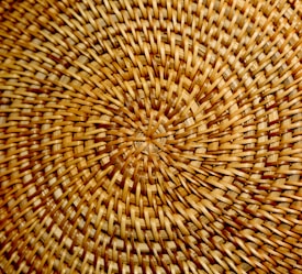 A close-up view of a woven basket pattern displaying intricate, circular weaving with various shades of brown and beige. The texture is smooth with a natural and rustic appearance.