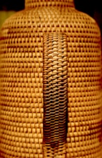 A close-up view of a woven wicker texture with a central handle. The weaving pattern is intricate, featuring interlaced strands of material that create a uniform, textured surface.