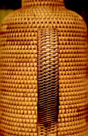 A close-up view of a woven wicker texture with a central handle. The weaving pattern is intricate, featuring interlaced strands of material that create a uniform, textured surface.