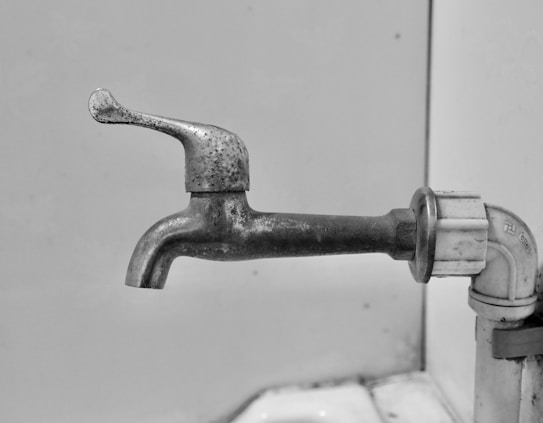 An old, metal faucet with a slightly rough and tarnished surface, emerging from a white pipe. The faucet handle has a simple design and is turned to the side. The background is a plain light-colored wall with some minor stains.