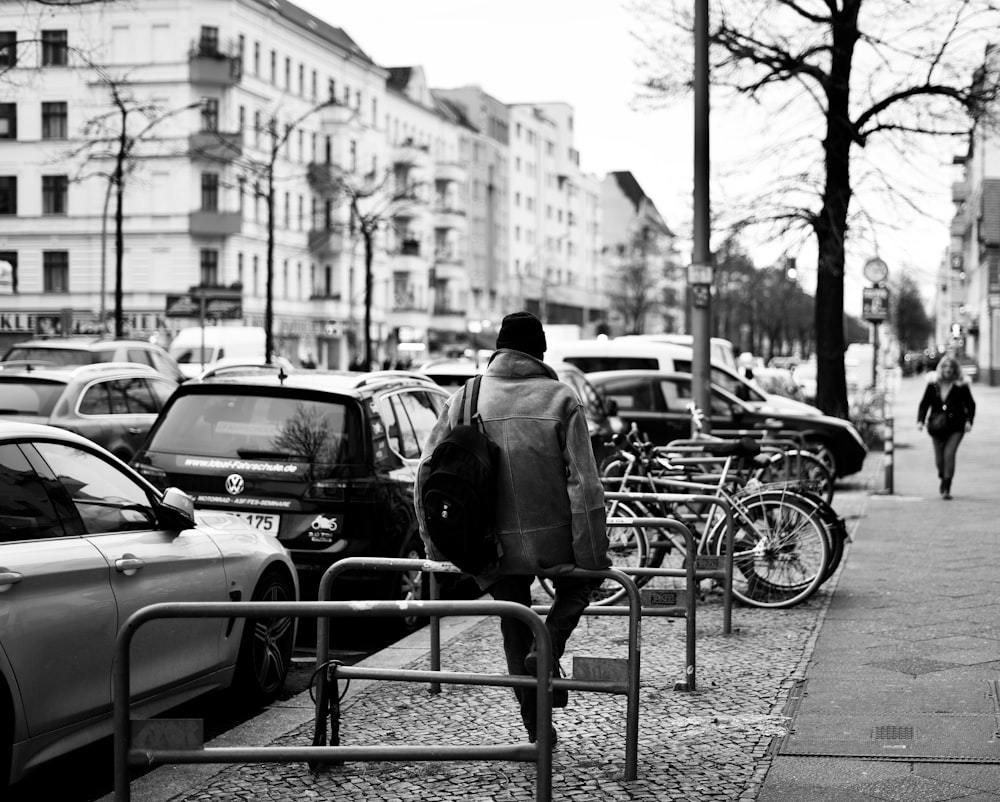 grayscale photo of man in black jacket walking on sidewalk