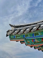 A traditional architectural roof with intricate designs is depicted, featuring colorful patterns in green, blue, red, and black. The roof is adorned with decorative tiles and carvings, all set against a backdrop of a cloudy sky.
