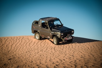A rugged off-road vehicle is positioned atop a sand dune under a clear blue sky. The vehicle is a compact four-wheel-drive with noticeable off-road tires and utility equipment attached, indicating readiness for desert terrain.