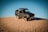 A rugged off-road vehicle is positioned atop a sand dune under a clear blue sky. The vehicle is a compact four-wheel-drive with noticeable off-road tires and utility equipment attached, indicating readiness for desert terrain.
