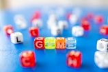 Colorful lettered dice are scattered on a blue surface, with the word 'GAME' spelled out prominently in the foreground using transparent dice. The dice are primarily red, orange, yellow, and clear, and there are other lettered dice visible in the background, creating a playful and vibrant scene.