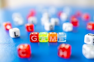 Colorful lettered dice are scattered on a blue surface, with the word 'GAME' spelled out prominently in the foreground using transparent dice. The dice are primarily red, orange, yellow, and clear, and there are other lettered dice visible in the background, creating a playful and vibrant scene.