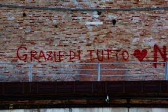 Red graffiti on a brick wall spells 'GRAZIE DI TUTTO' with a heart symbol and the letter 'N'. A metal railing runs horizontally across the wall below the graffiti.