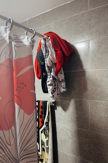 Several pieces of clothing are hanging on a hook attached to a tiled wall. A shower curtain with a floral design featuring red and white colors is hanging on a rod nearby. Below, a colorful towel with black, red, white, and green patterns is draped over another hook.