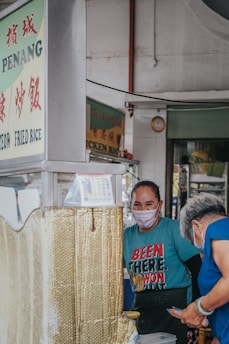 A street food vendor wearing a mask, standing at a stall with signage in a mix of English and another language. The vendor is interacting with an older woman who is also wearing a mask. The vendor wears a bright blue t-shirt with bold text, and both appear to be engaged in a friendly exchange.