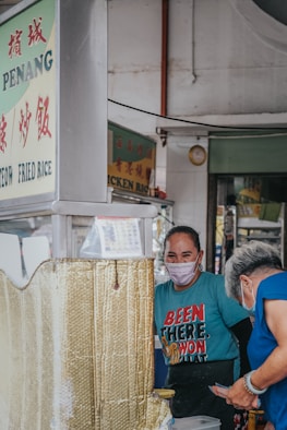 A street food vendor wearing a mask, standing at a stall with signage in a mix of English and another language. The vendor is interacting with an older woman who is also wearing a mask. The vendor wears a bright blue t-shirt with bold text, and both appear to be engaged in a friendly exchange.