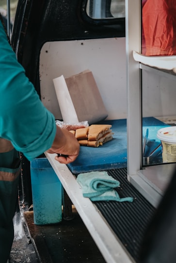 A person in a green shirt is preparing sandwiches on a blue cutting board inside a vehicle. There is a roll of bread being cut, a paper bag, and a tub of spreadable cream or butter nearby. A turquoise cloth is placed on the surface.