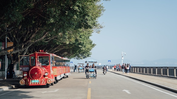 A red tourist train is parked along a tree-lined road near a waterfront. People are walking, riding bicycles, and enjoying the scenic view. The walkway is shaded by large trees on one side and bordered by a railing on the other. The sky is clear and the surrounding environment is bright and lively.