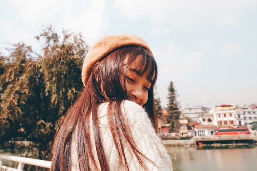 A smiling young woman wearing a pastel pink beret styled with a cozy sweater in a sunlit urban park.