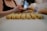A person is preparing or arranging small cookie-like pastries on a baking tray lined with parchment paper. The pastries are neatly aligned in rows, showing a textured surface and a light golden color. The focus is on the pastries, with the person's blurred figure in the background.