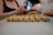A person is preparing or arranging small cookie-like pastries on a baking tray lined with parchment paper. The pastries are neatly aligned in rows, showing a textured surface and a light golden color. The focus is on the pastries, with the person's blurred figure in the background.