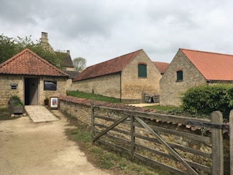 A rustic scene with several stone buildings featuring red-tiled roofs, surrounded by a wooden fence and lush greenery. The foreground includes a small building marked as the entrance to a museum, bordered by a stone path and a decorative arrangement of plants.