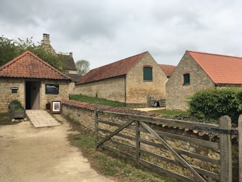 A rustic scene with several stone buildings featuring red-tiled roofs, surrounded by a wooden fence and lush greenery. The foreground includes a small building marked as the entrance to a museum, bordered by a stone path and a decorative arrangement of plants.