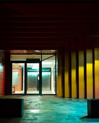 A modern building entrance with glass doors and visible elevators inside. The walls are adorned with a combination of red and yellow colors, and the interior is brightly lit. The exterior features a textured stone floor and muted lighting, which creates a contrasting atmosphere.