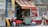 A small street food stall with a red and yellow striped awning displays the name 'Tortilleria La Palma.' A person wearing a purple jacket and red cap sits at a table with a red tablecloth, while another person kneels nearby in front of a Coca-Cola sign. The surroundings include a closed metal shutter with painted text and a man walking by carrying bags.