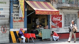 A small street food stall with a red and yellow striped awning displays the name 'Tortilleria La Palma.' A person wearing a purple jacket and red cap sits at a table with a red tablecloth, while another person kneels nearby in front of a Coca-Cola sign. The surroundings include a closed metal shutter with painted text and a man walking by carrying bags.
