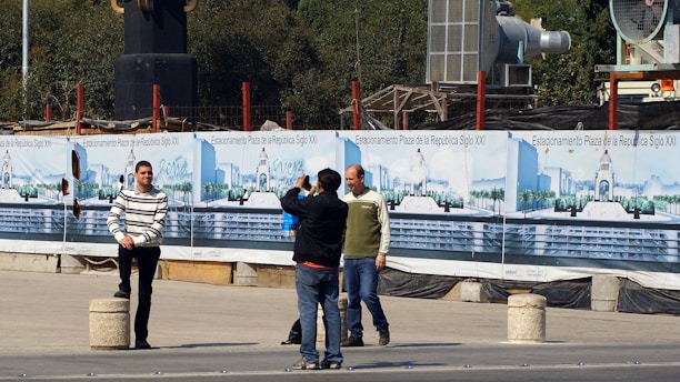 Several people are in an outdoor area near a construction site. One person in a striped sweater poses for a photograph while another holds a camera. Behind them are banners displaying images and text related to 'Estacionamiento Plaza de la República Siglo XXI'. Construction materials and equipment are visible in the background.