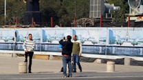 Several people are in an outdoor area near a construction site. One person in a striped sweater poses for a photograph while another holds a camera. Behind them are banners displaying images and text related to 'Estacionamiento Plaza de la Rep&uacute;blica Siglo XXI'. Construction materials and equipment are visible in the background.