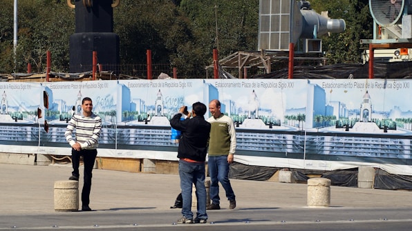 Several people are in an outdoor area near a construction site. One person in a striped sweater poses for a photograph while another holds a camera. Behind them are banners displaying images and text related to 'Estacionamiento Plaza de la República Siglo XXI'. Construction materials and equipment are visible in the background.