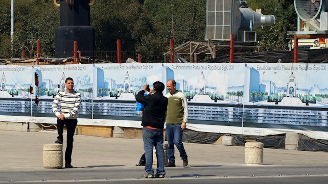 Several people are in an outdoor area near a construction site. One person in a striped sweater poses for a photograph while another holds a camera. Behind them are banners displaying images and text related to 'Estacionamiento Plaza de la Rep&uacute;blica Siglo XXI'. Construction materials and equipment are visible in the background.