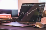 A laptop displaying a stock market graph with a rising trend is placed on a desk. Next to the laptop are several books stacked on each other, with titles related to investing. A mouse is positioned to the right of the laptop.