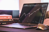A laptop displaying a stock market graph with a rising trend is placed on a desk. Next to the laptop are several books stacked on each other, with titles related to investing. A mouse is positioned to the right of the laptop.